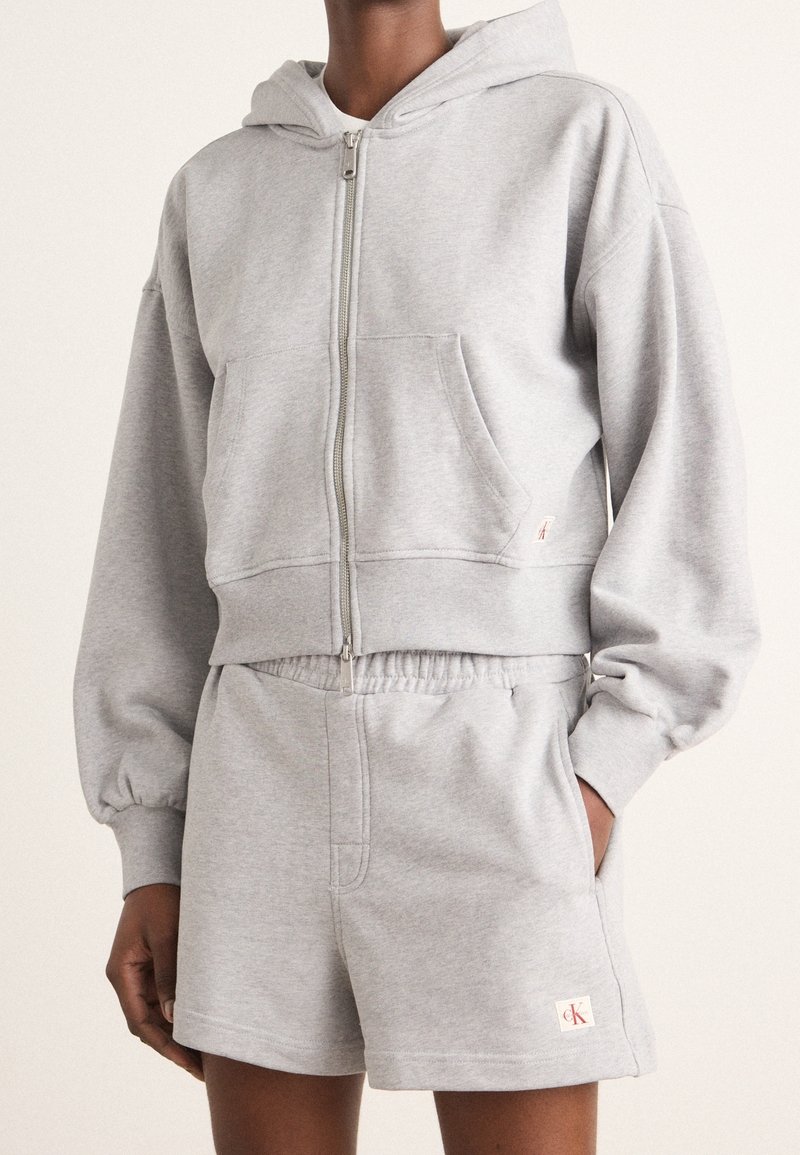 Sudadera gris con cremallera y bolsillo frontal, combinada con pantalones cortos grises a juego que cuentan con un parche de logo y cinturilla elástica.