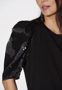 Schwarzes Oberteil mit transparenten, glänzenden schwarzen Puffärmeln, die an den Nähten mit kleinen Strasssteinen verziert sind. Weicher Stoff und lockere Passform.