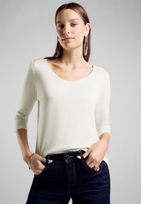 Weißes Oberteil mit langen Ärmeln und weitem Ausschnitt, kombiniert mit dunkelblauen Jeans; auffälliger Ring und Ohrringe als Accessoires. Glatte Textur.