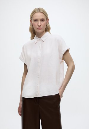 Frau mit langen blonden Haaren, die ein weißes, kurzärmliges Hemd mit Knöpfen und dunkelbraune Hose trägt, steht mit einer Hand in der Tasche.