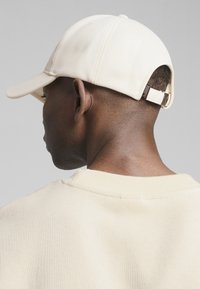 Beige pet met een gebogen klep, gemaakt van gestructureerde stof. Verstelbare band aan de achterkant. Draagt een licht beige geribbeld trui.