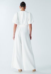 Witte jumpsuit met wijde pijpen, korte pofmouwen en een getailleerde taille. Gladde stof met een ritsdetail aan de achterkant. Draagt nude hoge hakken.