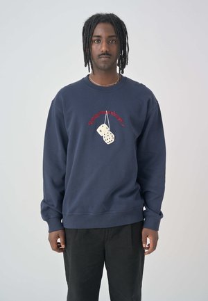Navy sweatshirt with front embroidery featuring two dice. Comfortable fit, ribbed cuffs, and hem. Worn with black pants.