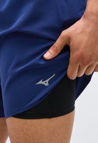 Χέρι τραβάει προς τα πάνω σκούρο μπλε σορτς για να αποκαλύψει μαύρα σορτς συμπίεσης με αντανακλαστικό λογότυπο στο πάνω μέρος του μηρού.