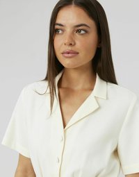 Jeune femme avec de longs cheveux bruns portant une chemise courte à manches courtes crème, regardant légèrement vers sa droite sur un fond uni.