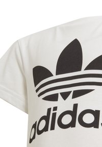 Bijela pamučna majica s velikim crnim Adidas logotipom i tri horizontalne pruge. Kratki rukavi i dizajn s okruglim izrezom.