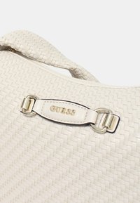 Borsa a mano intrecciata bianca con texture liscia, caratterizzata da un fermaglio in metallo argentato e logo "GUESS" su un'etichetta in pelle. Design con manico curvo.