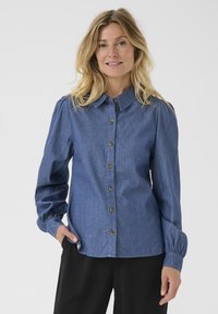 Chemise en denim à boutons avec manches longues, col arrondi et boutons marron. Présente une couleur bleu clair et des poignets de manches froncés.