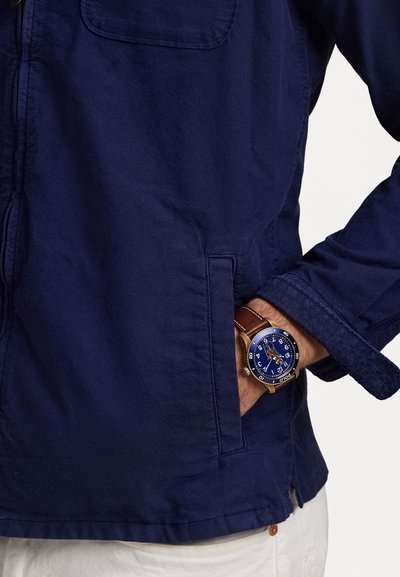 Chaqueta azul marino con un bolsillo en el pecho, que se lleva con un reloj de pulsera de cuero marrón que presenta una esfera azul y marcadores de hora blancos en contraste.