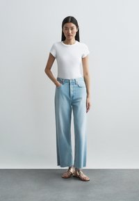 Valkoinen verkkokudottu lyhythihainen paita yhdistettynä korkeavyötäröisiin vaaleansinsisiin leveälahkeisiin farkkuihin. Huomionarvoinen repsottava helma; mallilla on kultaiset sandaalit.