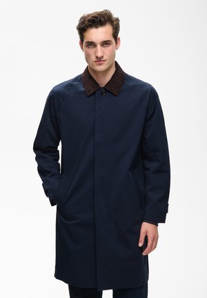 Jeune homme aux cheveux courts bruns portant un manteau bleu foncé avec un col noir, debout avec une main dans la poche, sur fond blanc.