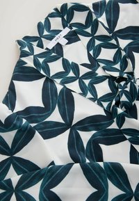 Tissu blanc avec un motif de feuilles géométriques vert foncé et une étiquette de vêtement indiquant « NEXT » près de la zone du col.