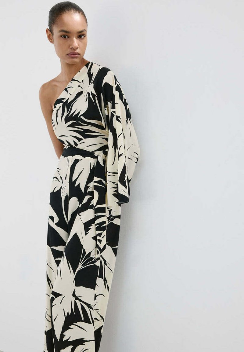 Femme portant une combinaison noire et blanche une épaule avec un grand motif de feuilles, se tenant devant un fond gris clair uni.