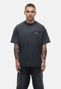 Krótki rękaw, ciemnoszara bawełniana koszulka z okrągłym dekoltem, z nadrukowanym logo na lewej piersi i luźnym krojem. Subtelna faktura widoczna.