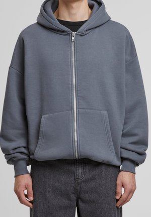 Osoba nosząca szarą bluzę rozpinaną na zamek z przednimi kieszeniami i ściągaczami, na czarnym t-shircie, w połączeniu z ciemnoszarymi jeansami.