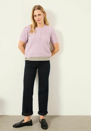 Lilac knitted short-sleeve top with ribbed hem, paired with dark denim jeans rolled at the cuff, and black loafers on a textured surface.