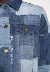 Chaqueta de mezclilla patchwork con diferentes tonos de azul, bordes deshilachados y botones metálicos grabados con "Michael Kors" en el bolsillo del pecho y en la parte frontal.