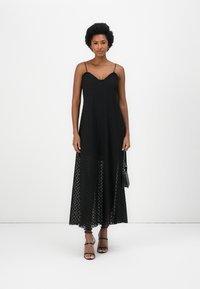 Black maxi dress with textured fabric, V-neckline, and thin adjustable straps. Sheer lower layer with a subtle pattern, paired with black heels.