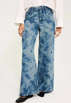 Højtaljede viddebenede jeans med blå tie-dye mønster, båret med sorte flade sko og en hvid langærmet skjorte.