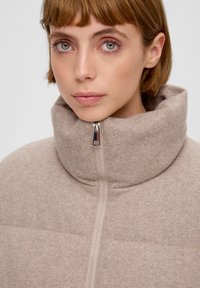 Beżowa kurtka z wysokim kołnierzem, wykonana z teksturowanej tkaniny, z pełnym zamkiem błyskawicznym i subtelnymi poziomymi szwami. Miękka w wyglądzie z minimalnymi akcentami.