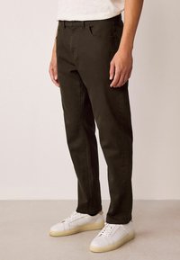 Tumedoolivroheline puuvillane püksid sirge lõikega, viie taskuga ja vöörihmadega, koos valgete madala kontsaga tossudega.