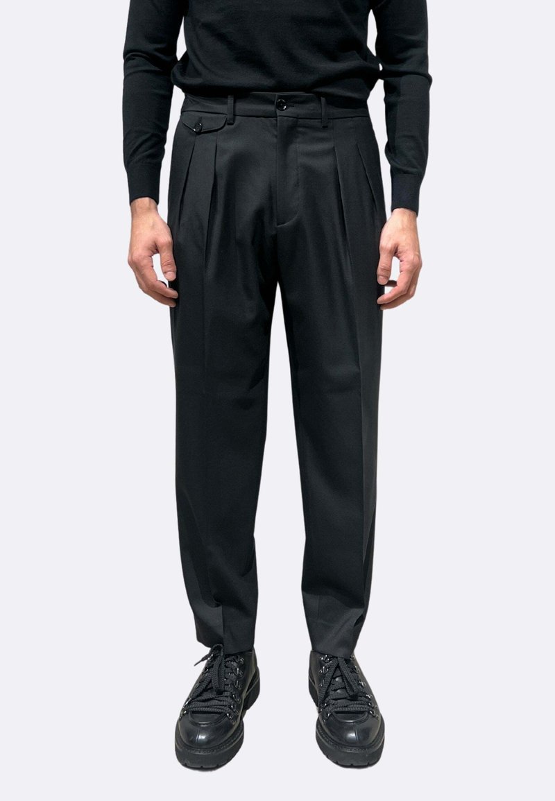 Pantaloni neri a vita alta con pieghe frontali, tasche laterali e una fascia spessa in vita, abbinati a scarpe nere stringate.