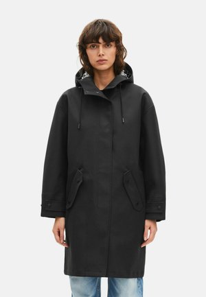 Kvinde iført sort hættejakke med lommer foran og manchetter med knapper, kombineret med lyseblå jeans, stående mod en hvid baggrund.