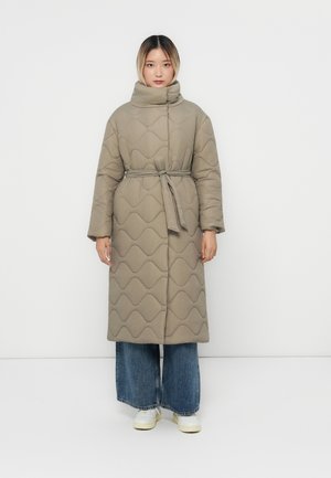 Une femme se tient face à l'objectif, portant un long manteau matelassé beige avec une ceinture, un jean bleu ample et des baskets blanches, le tout devant un fond uni.