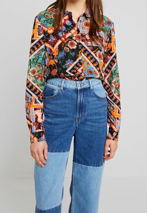 Kvinde iført knaplukket farverig blomstermønstret bluse, stoppet ned i to-tone blå lap-jeans, stående mod en ensfarvet baggrund.
