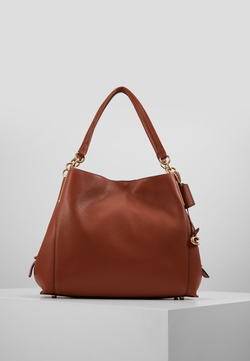 Sac à main en cuir grainé marron avec une bandoulière large unique, une quincaillerie dorée et une petite étiquette en cuir attachée sur un socle gris.