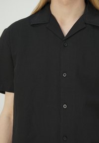 Personne portant une chemise noire à manches courtes avec col, ne montrant que la poitrine et la zone du cou.