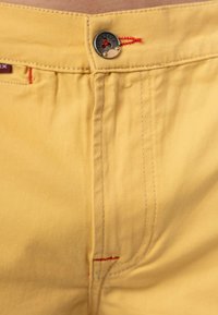 Shorts gialli in cotone con cuciture rosse, un bottone in metallo argentato e due tasche frontali. Il tessuto è liscio con una leggera lucentezza.
