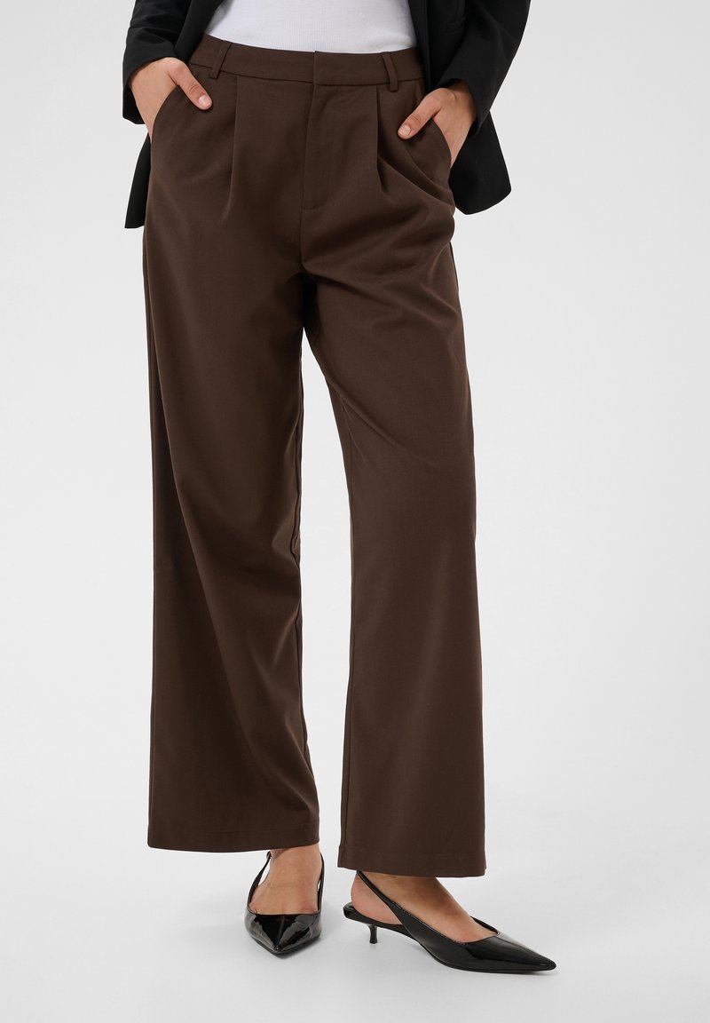 Personne portant un pantalon large marron foncé, des chaussures slingback noires à petit talon et bouts pointus, un top blanc et un blazer noir, les mains dans les poches.