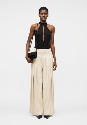 Nő fekete, ujjatlan, elöl kivágott pántos felsőben, széles, bézs pliszírozott nadrágban, fekete hegyes cipőben és fekete válltáskával.