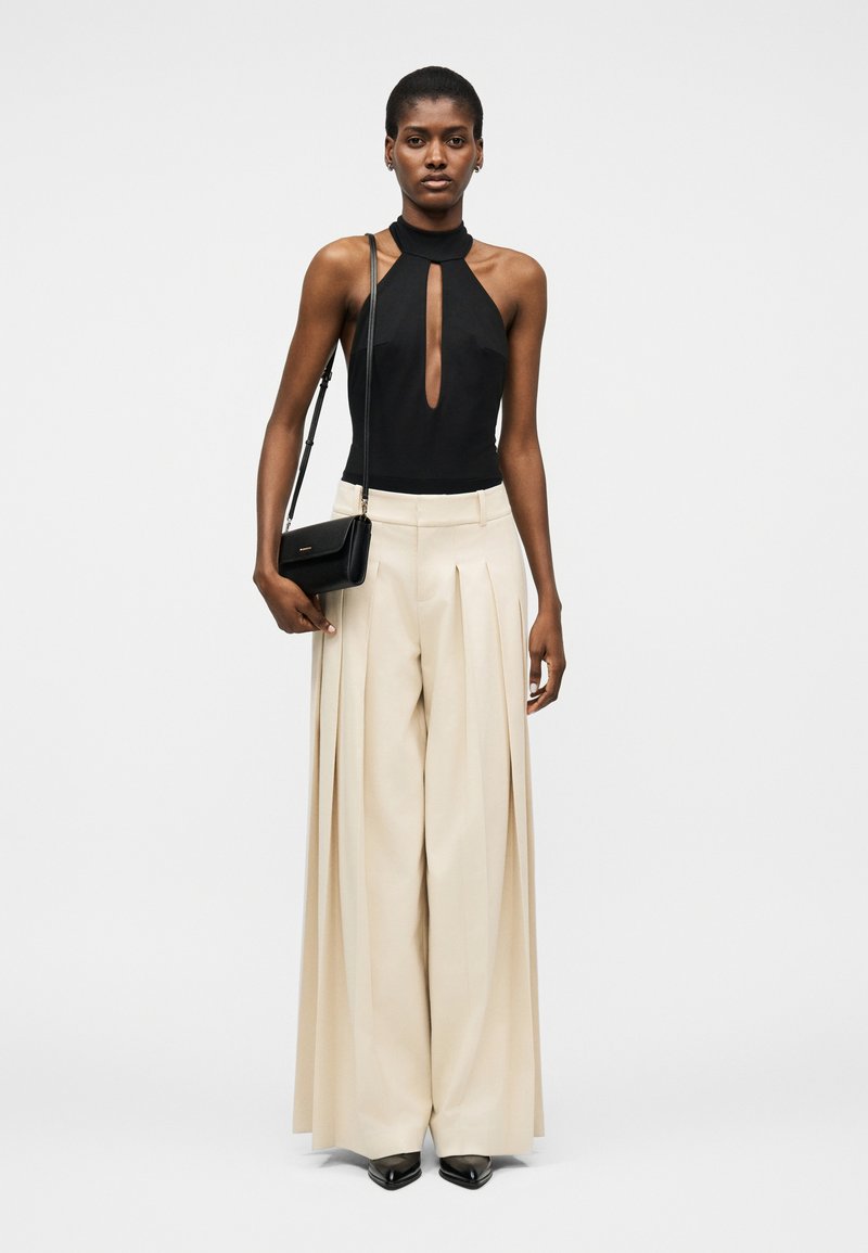 Γυναίκα που φοράει μαύρο τοπ χωρίς μανίκια με άνοιγμα στο μπροστινό μέρος, φαρδιές μπεζ πλισέ παντελόνες, μαύρα μυτερά παπούτσια και μαύρη τσάντα ώμου.