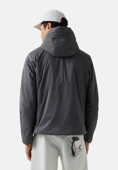 Man die van achteren kijkt, gekleed in een donkergrijze gewatteerde jas met capuchon, lichtgrijze broek, witte pet, met een zonnebril geklemd aan de broekzak.