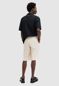 Maglietta nera a maniche corte con una vestibilità ampia, abbinata a pantaloni corti beige chiaro e scarpe nere con un dettaglio a strap. Vista posteriore su uno sfondo neutro.