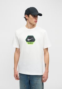 Νέος άνδρας με λευκό μπλουζάκι Nike με λογότυπο εξαγώνου και μαύρο καπέλο, κοιτάζει προς τα δεξιά, στέκεται μπροστά σε μονόχρωμο φωτεινό φόντο.