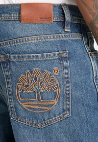 Jean en denim bleu avec logo Timberland en forme d'arbre brodé en marron sur la poche arrière et une étiquette en cuir Timberland sur la ceinture, montrant partiellement un bras tatoué.