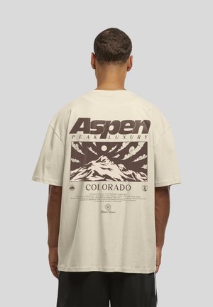 Homme de dos portant un t-shirt beige avec un graphisme marron de montagnes et un grand texte indiquant Aspen Peak Luxury Colorado.