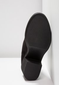 Semelle d'une botte à talon noire montrant une bande de roulement texturée et un talon bloc sur une surface blanche, avec un fond blanc.