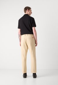 Polo noir avec col, associé à un pantalon beige et des chaussures noires. Le pantalon présente une poche arrière avec un petit détail en étiquette rouge.