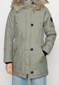 Parka giacca imbottita verde oliva con cappuccio foderato in pelliccia, chiusura a bottoni e zip, vita regolabile e due tasche frontali.