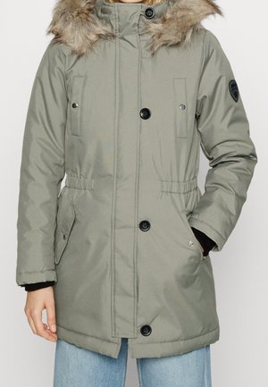 Parka acolchada de color verde oliva con capucha forrada de piel, cierre a presión y cremallera, cintura ajustable y dos bolsillos frontales.