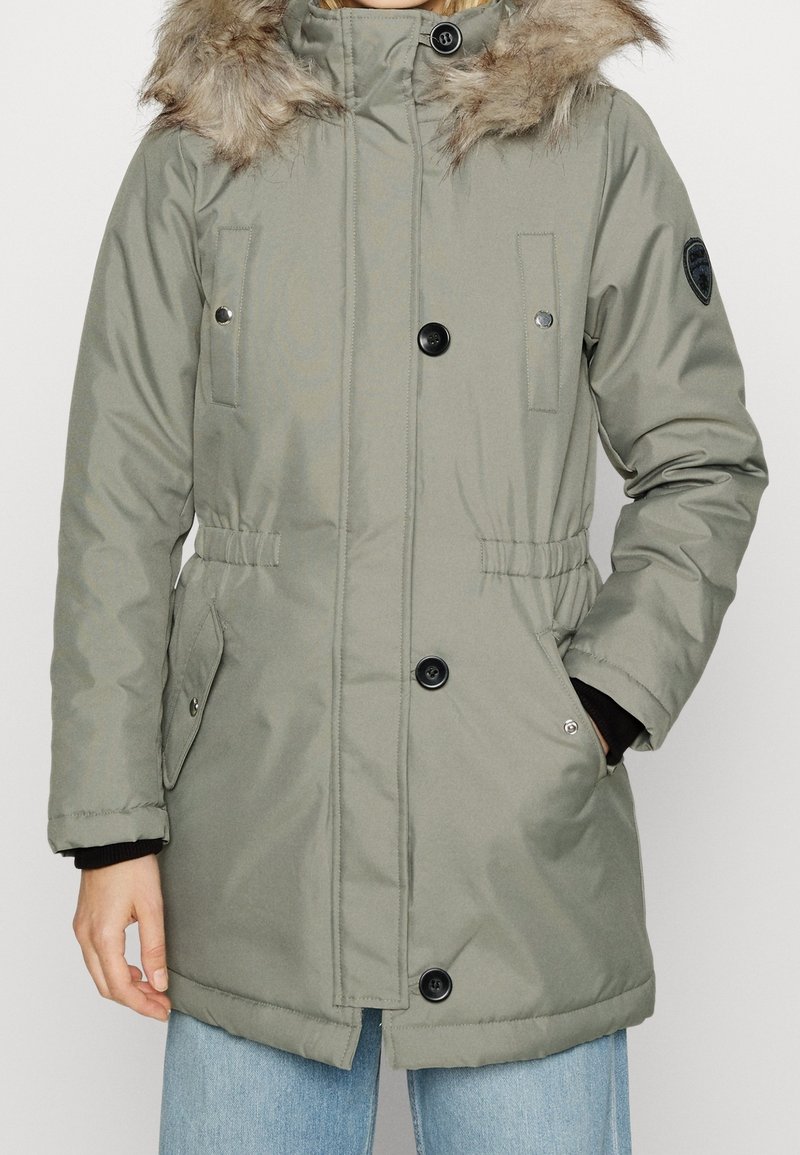 Parka giacca imbottita verde oliva con cappuccio foderato in pelliccia, chiusura a bottoni e zip, vita regolabile e due tasche frontali.