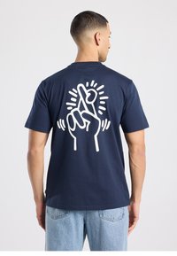 T-shirt en coton bleu marine avec un grand motif blanc d'une main croisant les doigts au dos, entouré de lignes rayonnantes autour du design.