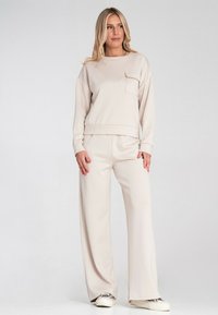 Beżowy zestaw odzieży wypoczynkowej składający się z długiego rękawa z kieszonką na piersi oraz spodni o szerokich nogawkach, wykonany z miękkiego materiału o gładkiej fakturze.