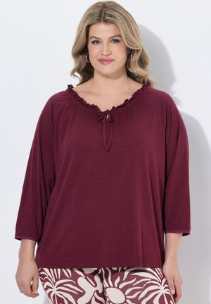 Donna con capelli biondi mossi che indossa una maglia bordeaux a maniche 3/4 con collo a fiocco e pantaloni con motivo a foglie bianche e bordeaux.