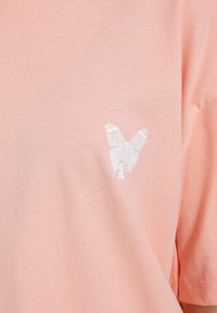 Camiseta de manga corta, de color rosa claro, hecha de algodón suave, que tiene un gráfico de mariposa impreso en blanco y texto en la parte frontal.