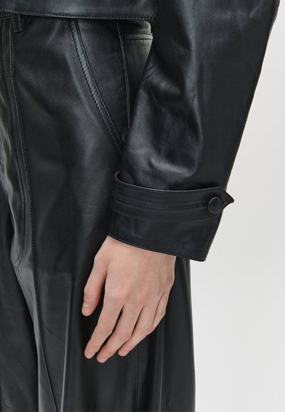 Približni pogled na ruku koja odmara sa strane, u crnoj kožnoj jakni s kopčanim manžetnama i usklađenim kožnim hlačama.